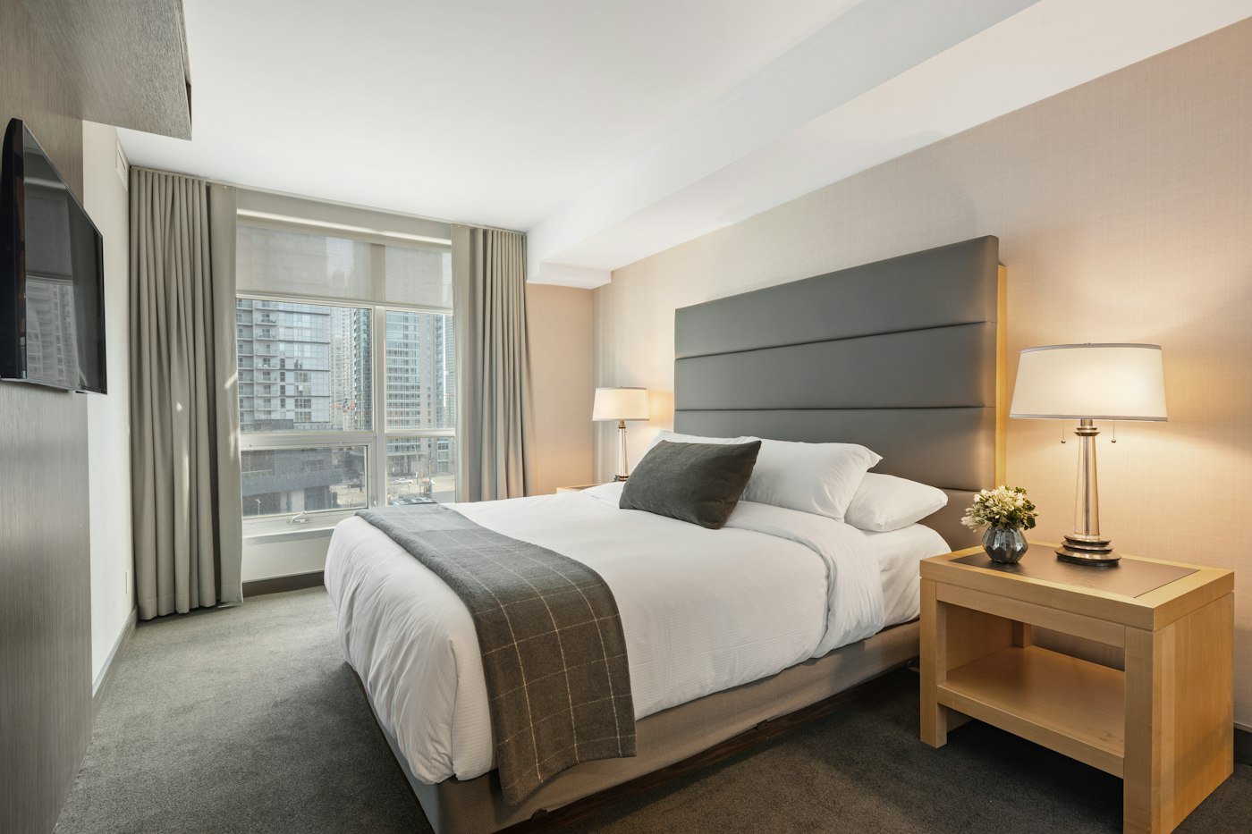 A white bed with a grey headboard, a wooden bedside table, and city buildings beyond the window